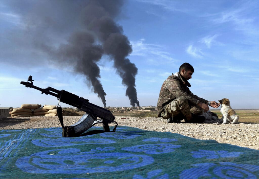 Des soldats kurdes syriens des YPG s'occupent de chiots à un poste de contrôle sur la route de Tel Tamir, en Syrie, le 12 novembre 2019. La fumée provenant de la combustion du pétrole est utilisée comme bouclier contre les drones et les frappes aériennes. Un conflit féroce a éclaté après le retrait des troupes du président américain Donald Trump, considéré par beaucoup comme une trahison des alliés kurdes © Carol Guzy