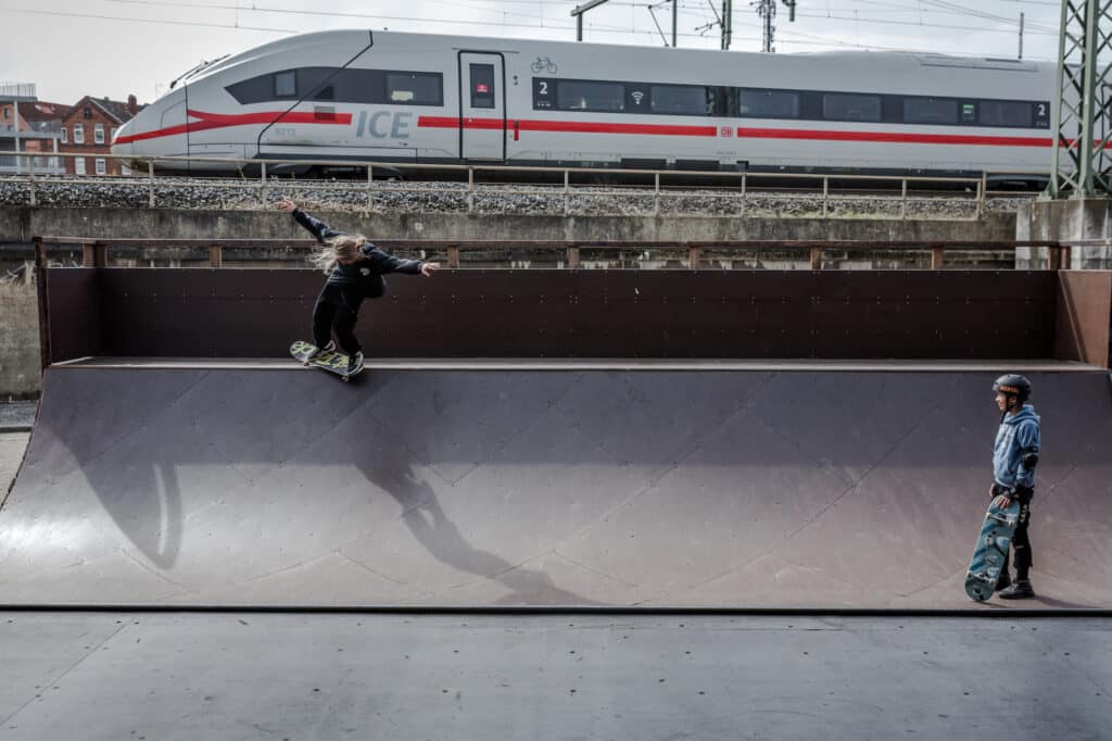 While a German train I.C.E (Intercity Express) passes-by the Gleis D Skate Halle, 12-year-old Abdullah stares at Yurii Korotun performing a trick on an outdoor mini halfpipe. Hanover, Germany. April 13th, 2022. © Thomas Girondel