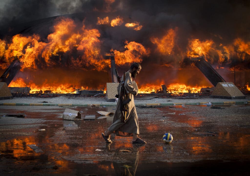 Tripoli, le 23 août 2011. Un homme, armé de deux kalachnikovs, pousse un ballon devant une tente incendiée du complexe de Bab al-Aziziya, le bumker de Kadhafi, qui vient de tomber aux mains des insurgés.