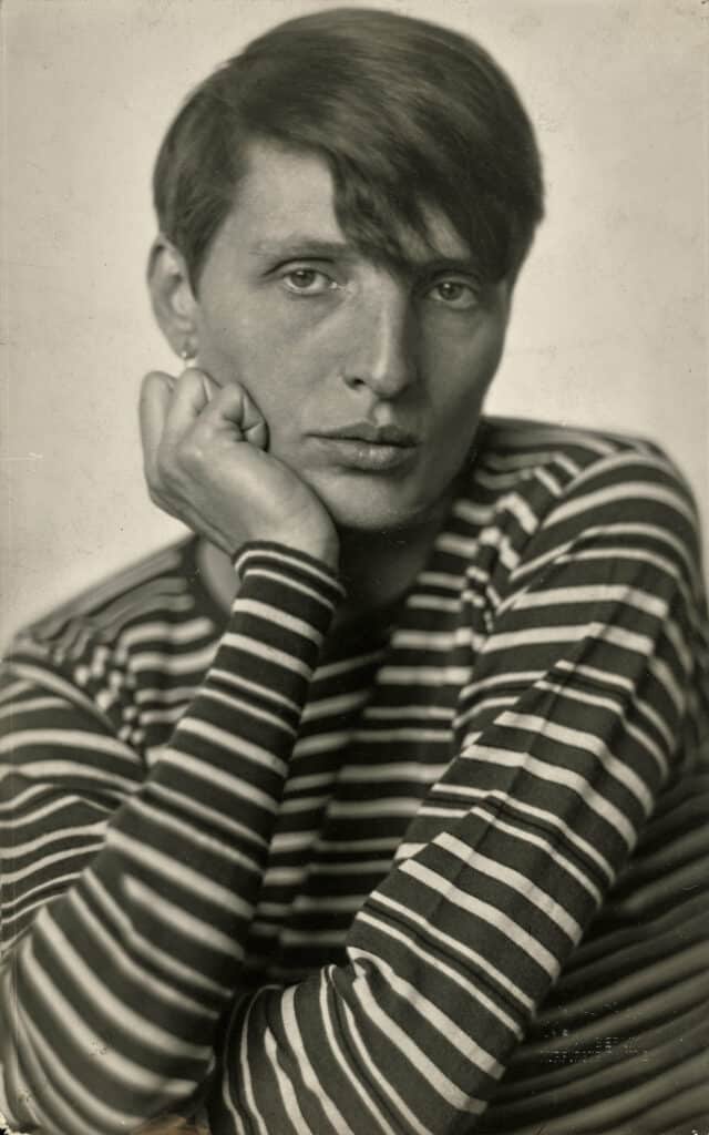 Portrait of Renee Sintenis, (Renate Alice Sintenis) German sculptor, wearing a long-sleeved striped top, with her face resting in her palm, while looking directly into the camera.