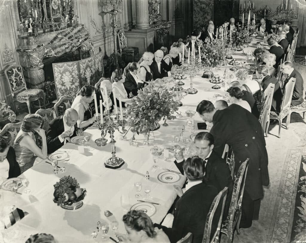 A formal dinner at the Vanderbilt house, hosted by Mrs. Vanderbilt, wearing one of her characteristic bandeaux at the center of the near side of the table set with silver settings for all forty-six guests