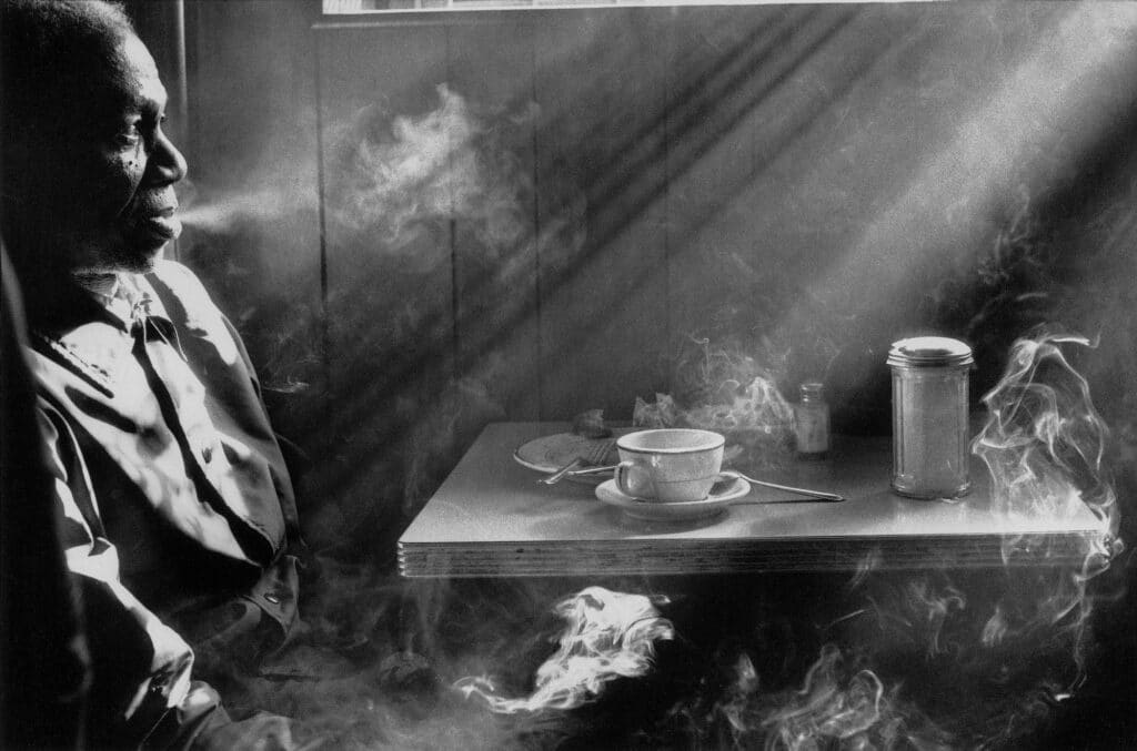 A man seated at a table in a diner exhales smoke while sunlight streams in the window, New York, 1974. © Harold Feinstein